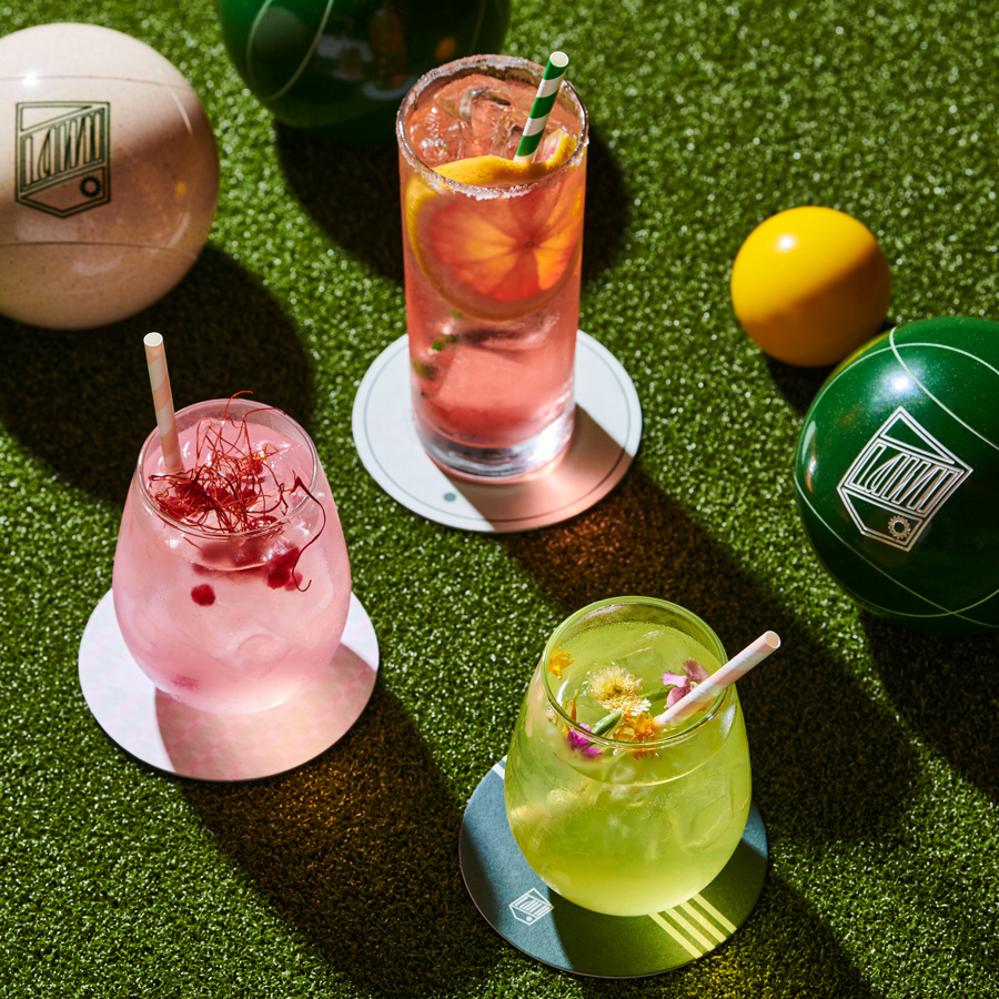 An overhead shot of three colorful cocktails on a faux grass surface, with lawn bowling balls scattered around. Each drink is uniquely garnished and presented on a small coaster with the establishment's logo. The sunlit setting suggests a relaxed, outdoor-inspired environment for leisure and socializing.