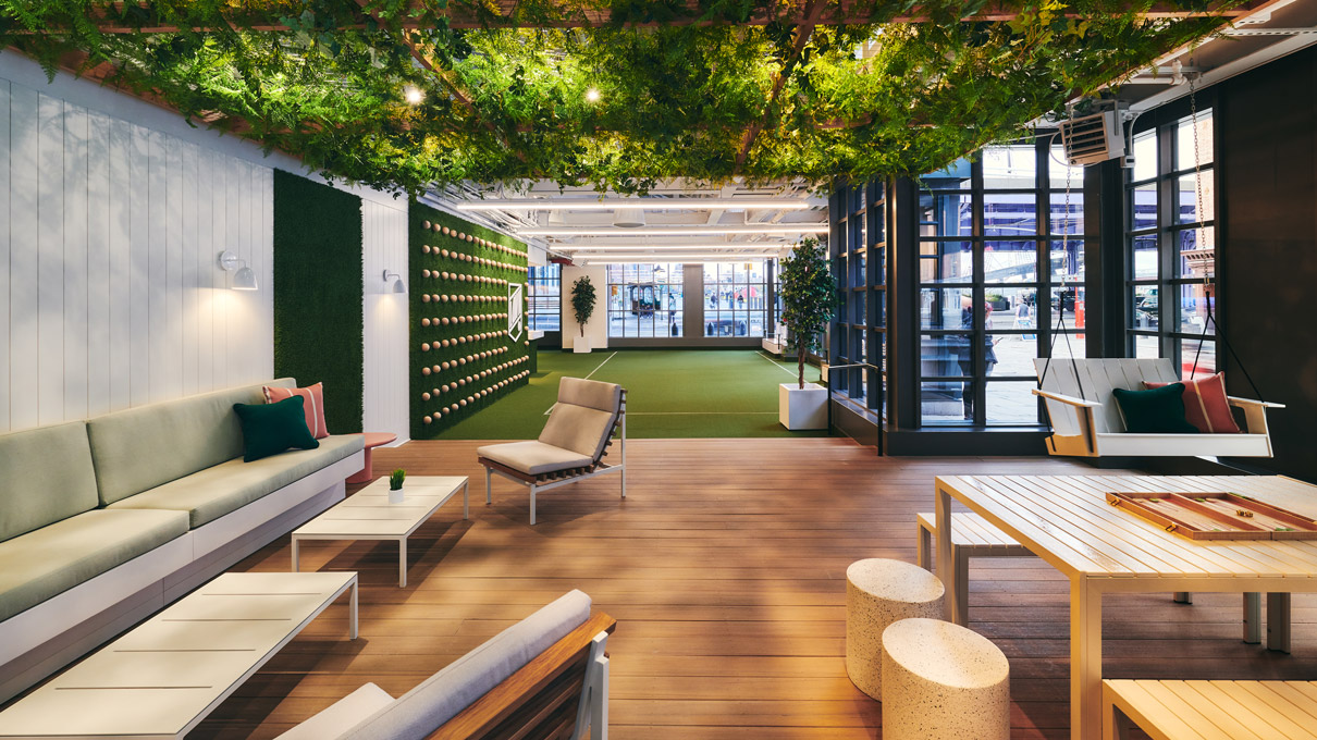 A modern indoor lounge area with a greenery ceiling, sleek benches, chairs, wooden tables, and large windows allowing natural light to accentuate the space's warm tones.