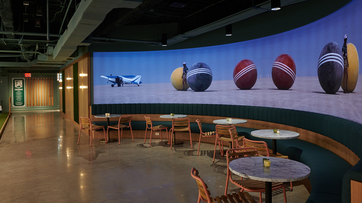 An indoor recreational space with a casual seating arrangement, round tables, and wooden chairs. A large mural of oversized lawn game balls and a paper airplane adorns the wall.
