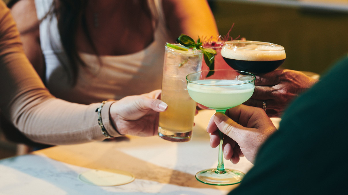 People in a social setting clinking glasses, with a focus on their hands and the colorful cocktails they're holding, highlighting a moment of celebration and camaraderie.