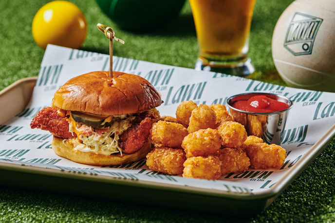 A delicious meal of a crispy chicken sandwich, tater tots, and a pint of beer on a tray with a green and yellow ball in the background, suggesting a fun, casual dining experience at a sports or entertainment venue.