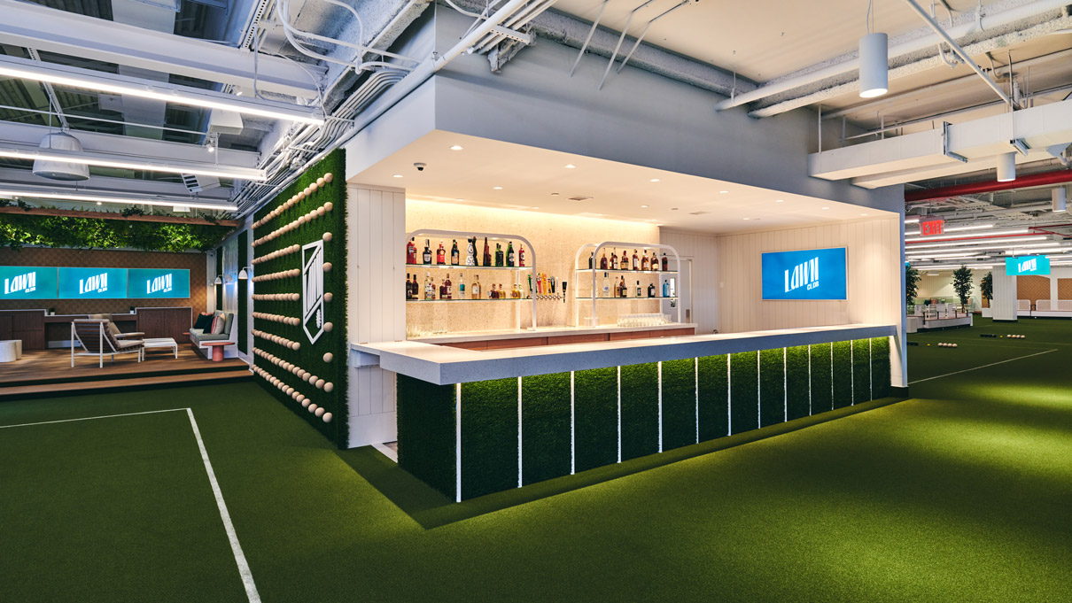 A contemporary indoor bar setup with a long counter and backlit shelves stocked with various bottles. The flooring is green astroturf, mimicking a grassy surface, and the space is brightly lit with overhead lights. Digital screens are visible in the background, enhancing the modern feel of the area.