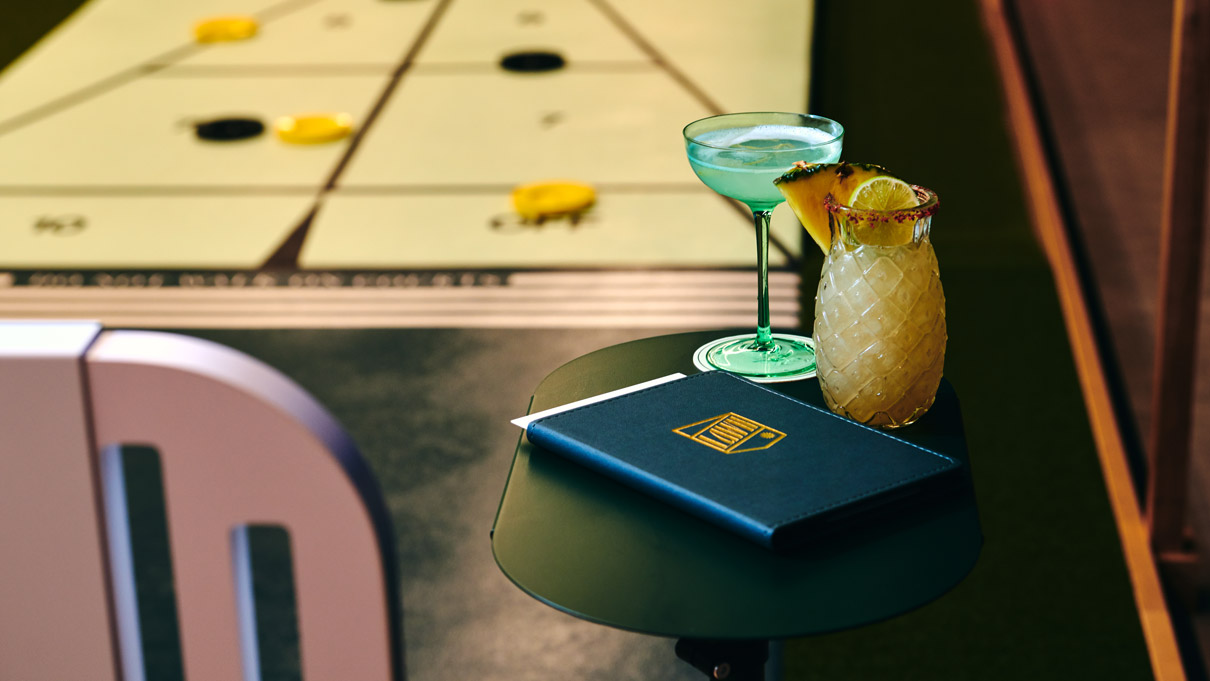 Two tropical cocktails on a small round table with a shuffleboard court in the background.