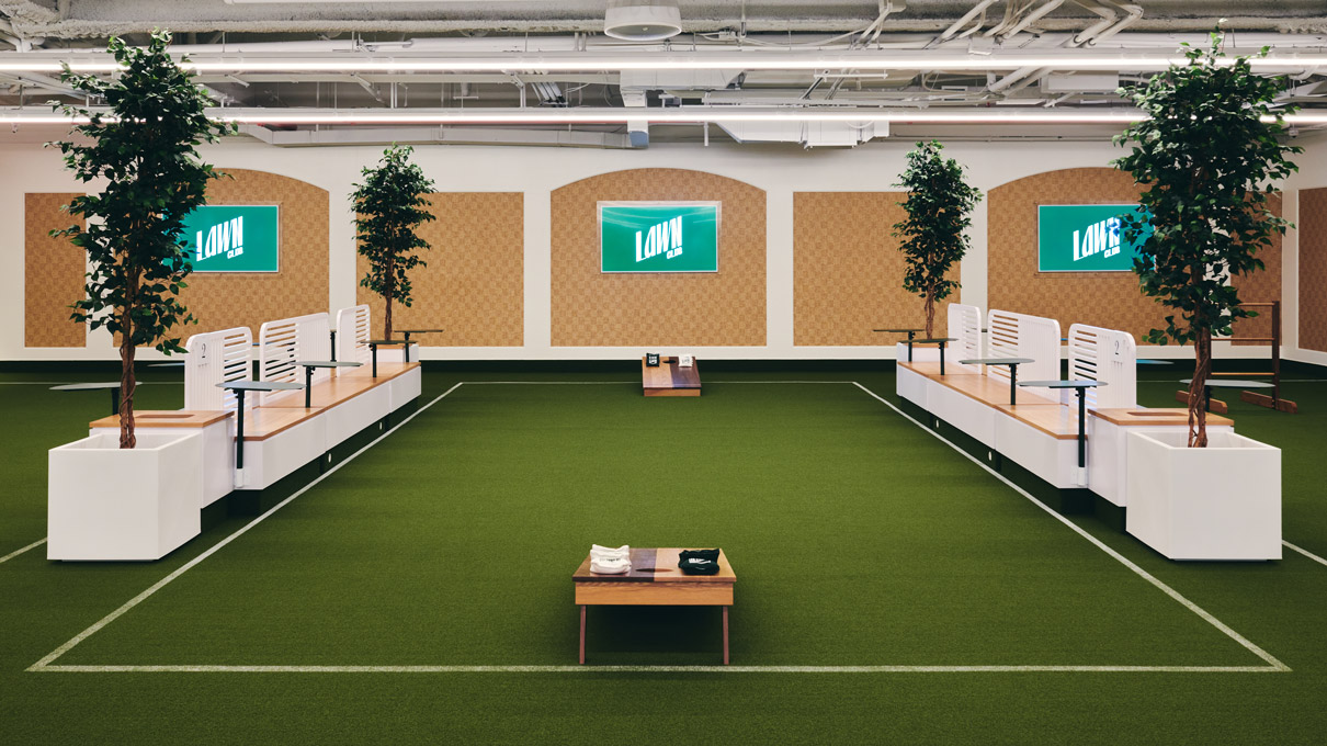 An indoor space creatively designed with an artificial turf floor, resembling a mini indoor park. The area is structured with white modular seating, each accompanied by a potted tree, enhancing the outdoor park feel. Three large monitors on the wall display the words “Lawn Club.”. The ceiling exposes modern industrial elements like piping and lighting, contrasting with the greenery and soft wood tones for a contemporary vibe.