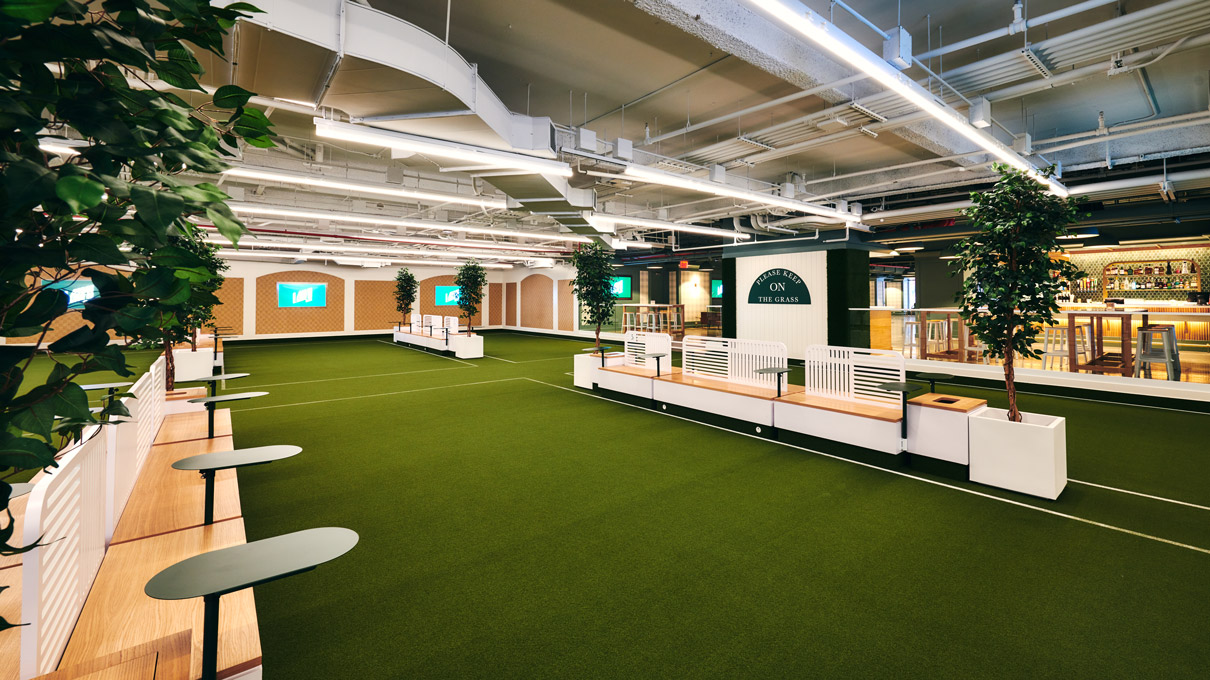 An indoor space creatively designed with an artificial turf floor, resembling a mini indoor park. The area is structured with white modular seating, each accompanied by a potted tree, enhancing the outdoor park feel. The ceiling exposes modern industrial elements like piping and lighting, contrasting with the greenery and soft wood tones for a contemporary vibe.