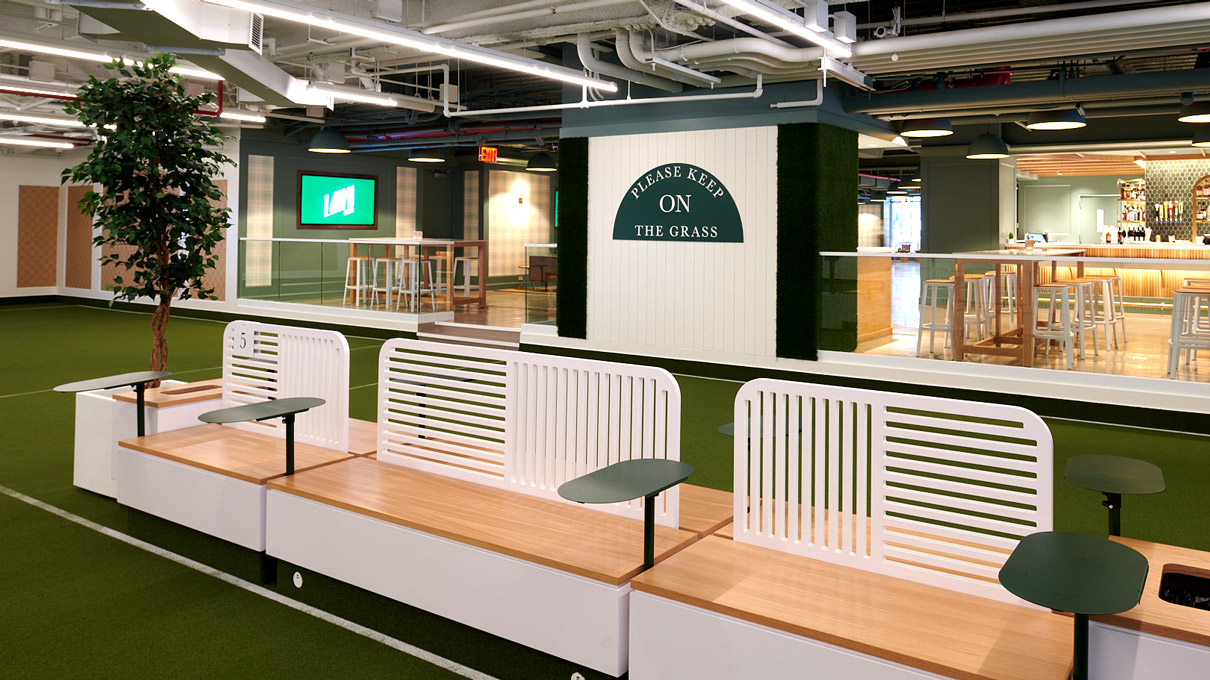 An indoor space creatively designed with an artificial turf floor, resembling a mini indoor park. The area is structured with white modular seating, each accompanied by a potted tree, enhancing the outdoor park feel. A sign in the background reads “Get on our lawn.” The ceiling exposes modern industrial elements like piping and lighting, contrasting with the greenery and soft wood tones for a contemporary vibe.