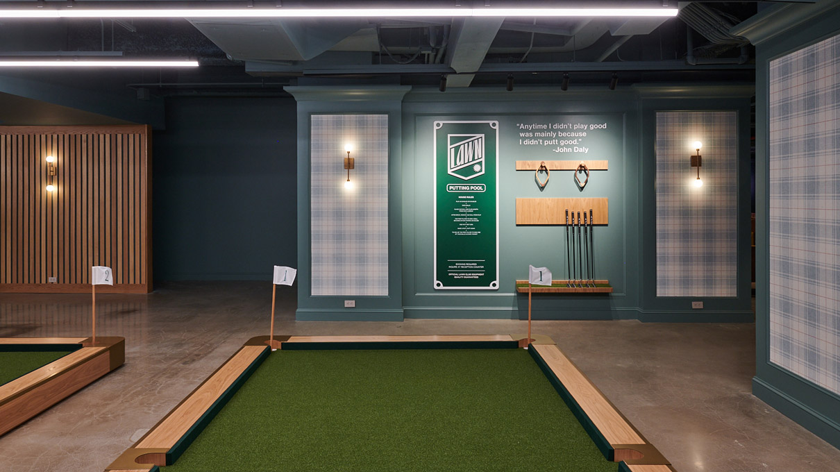 This is an image of an indoor recreational area with a modern design aesthetic. There's a golf putting green on the left, with two golf clubs ready for use. On the right, against a green wall, is a large infographic providing rules for a game. Above this, there is a minimalist wooden shelf displaying an array of golf balls, indicating a leisure or competitive aspect to the space.